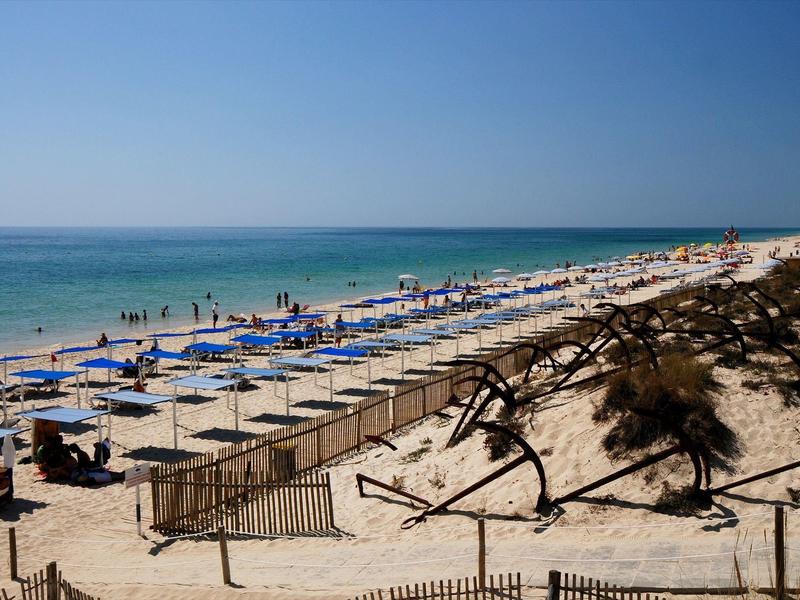 Playa con arena, sombrillas azules alineadas y mar tranquilo bajo cielo despejado.