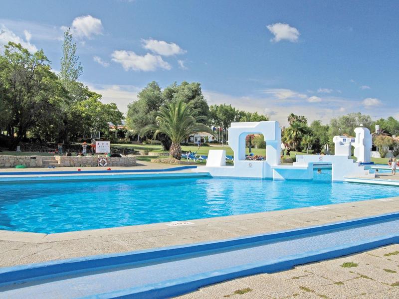Gran piscina azul con elementos arquitectónicos blancos y árboles al fondo bajo cielo azul.