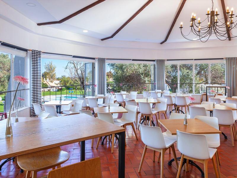 Comedor moderno y luminoso con sillas blancas, mesas de madera y grandes ventanas con vista al jardín.