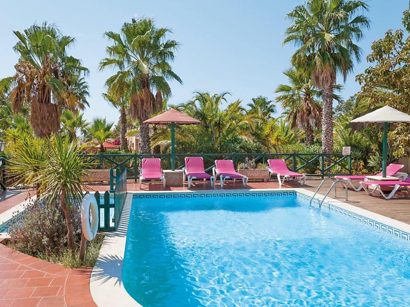 Außenpool mit klar blauem Wasser, rosa Liegestühlen, Palmen und Sonnenschirmen bei sonnigem Wetter.