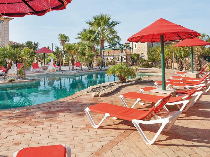 Poolbereich mit orangenen Liegestühlen und Sonnenschirmen, Palmen und klar blauem Himmel im Hintergrund.