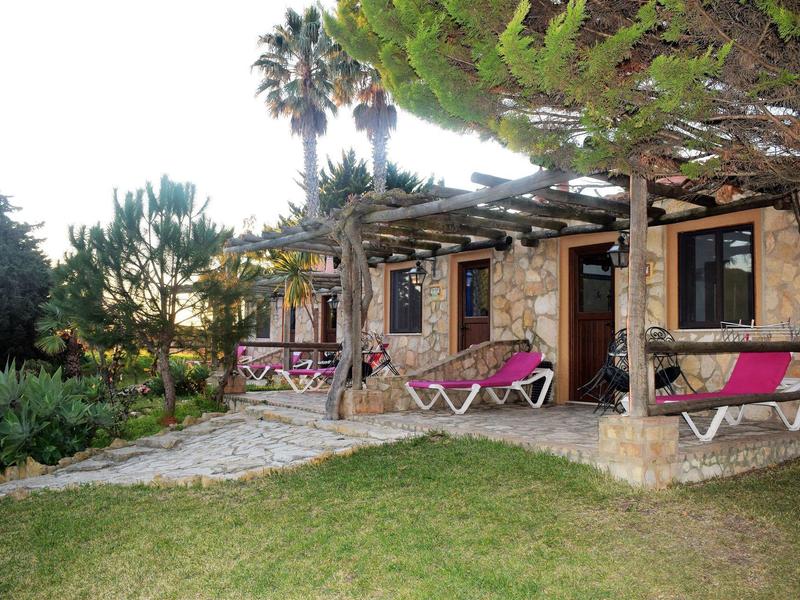 Rustikales Haus mit Steinmauer, Holzpergola, rosa Liegestühlen und grünem Garten davor.
