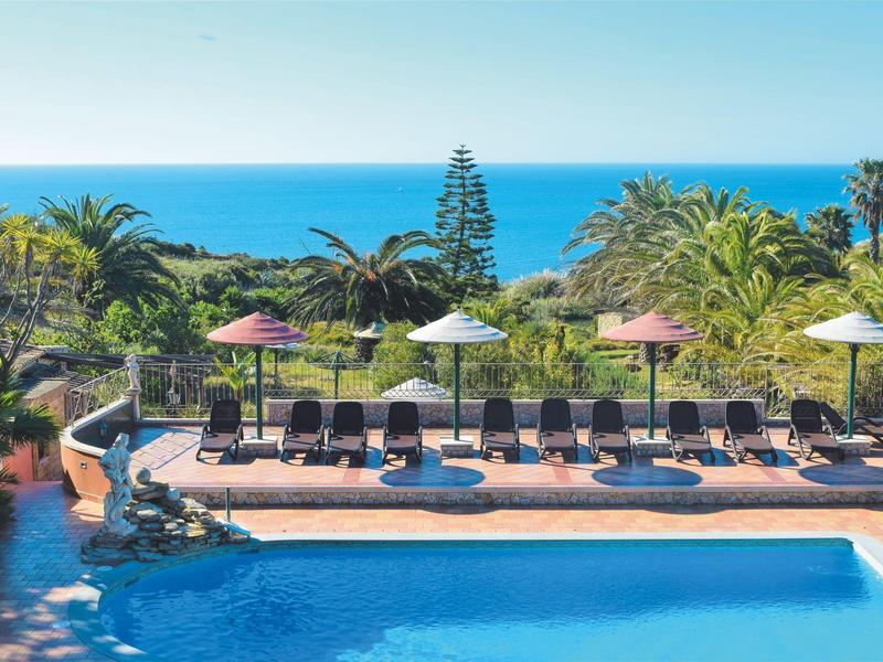 Großer Pool mit blauer Wasserfläche, Liegestühlen, Schirmen und Palmen vor blauem Meer im Hintergrund.