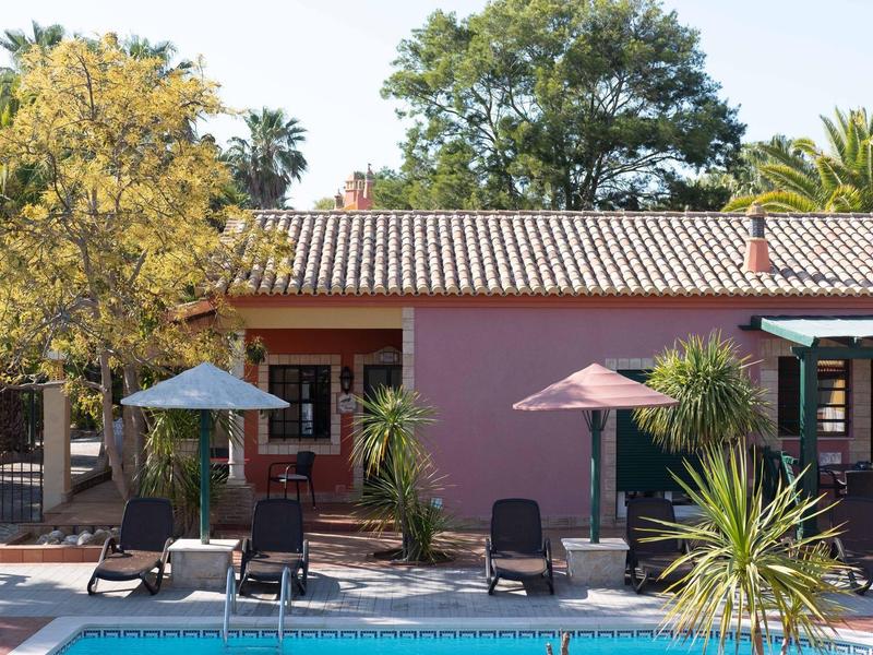 Ein rosa Haus neben einem Pool mit Liegestühlen, Sonnenschirmen und grünen Pflanzen.