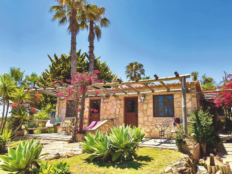 Kleines Steinhaus mit Pergola, Palmen und Blumen in sonniger, grüner Gartenoase.
