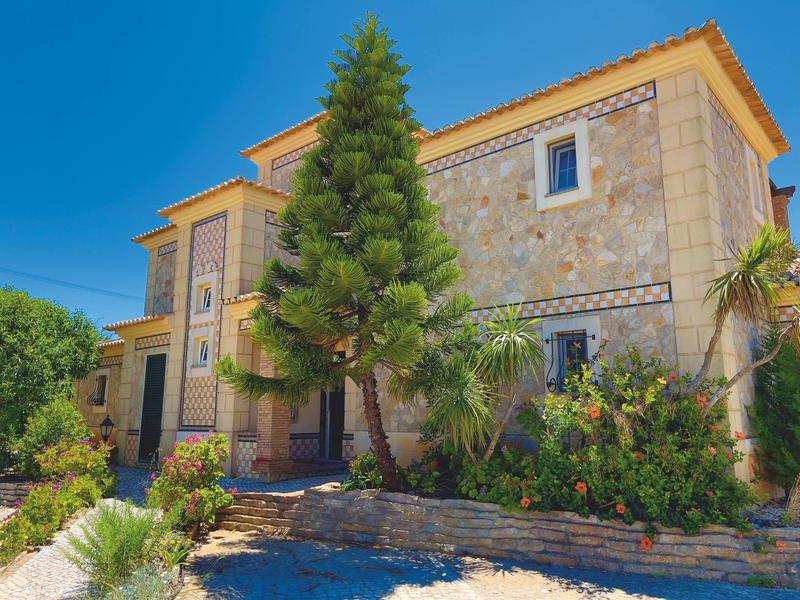 Steinernes zweistöckiges Haus mit orangenem Dach, umgeben von grünen Bäumen und Pflanzen.