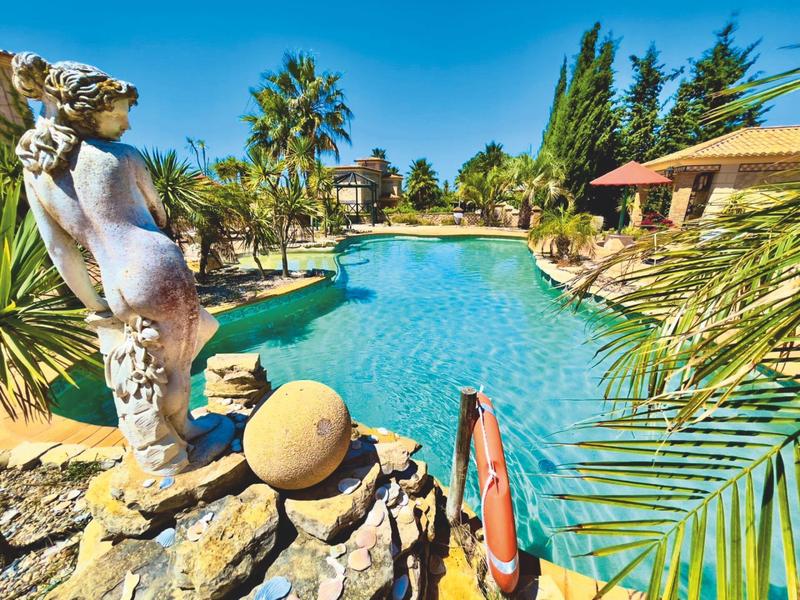 Großer Pool mit blauer Wasserfläche, Palmen, Steinskulptur und Sonnenschirmen unter blauem Himmel.