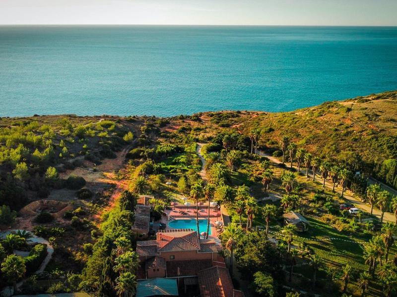Landschaft mit grün bewachsenem Hügel, Häusern und einem blauen Schwimmbad am blauen Meer im Hintergrund.