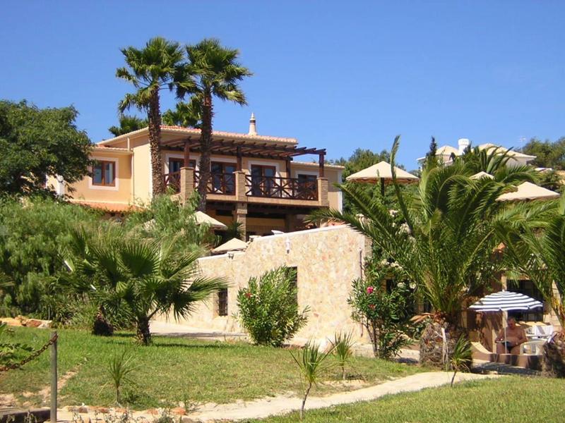Haus mit Terrasse, Palmen und grünem Garten unter blauem Himmel.
