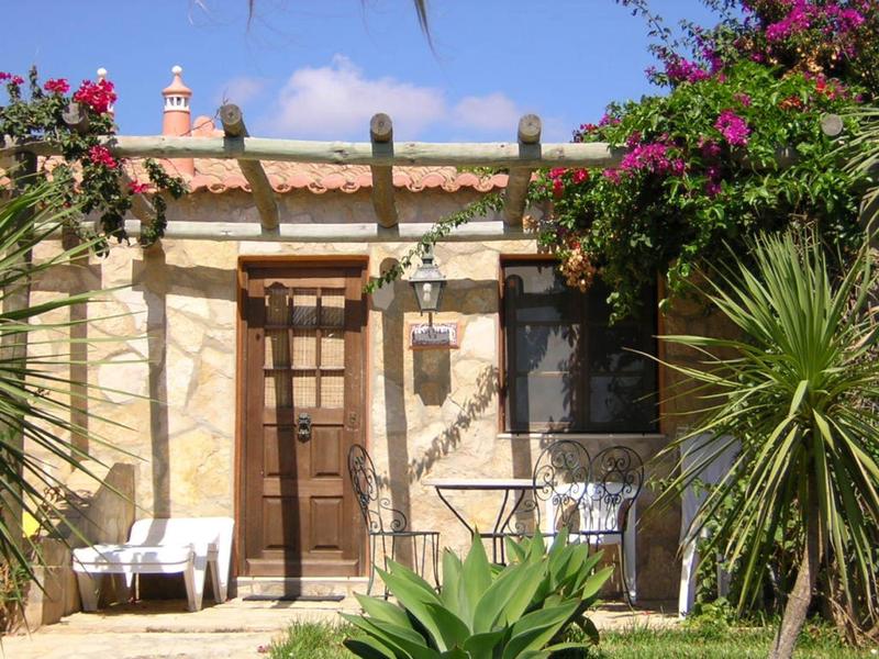 Kleines helles Haus mit braunem Holztor, weißen Stühlen, Pflanzen und blauem Himmel.
