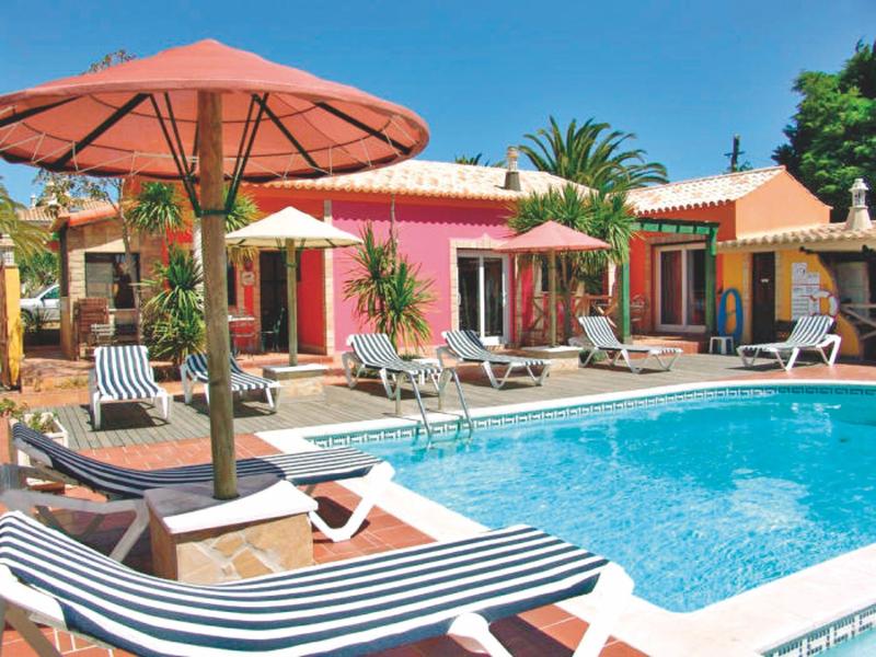 Farbenfrohes Haus mit Terrasse, mehreren Sonnenschirmen und Pool mit Liegestühlen am blauen Himmel.