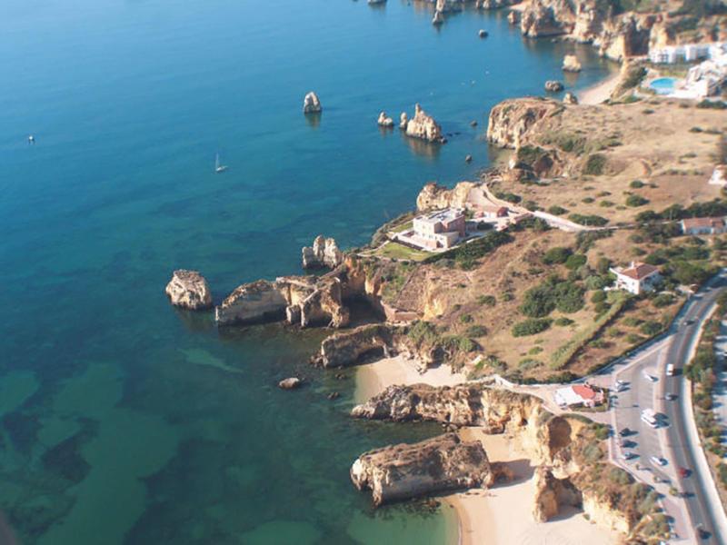 Küste mit Felsen, klarem blauem Wasser und einer Straße entlang felsigem Hang.
