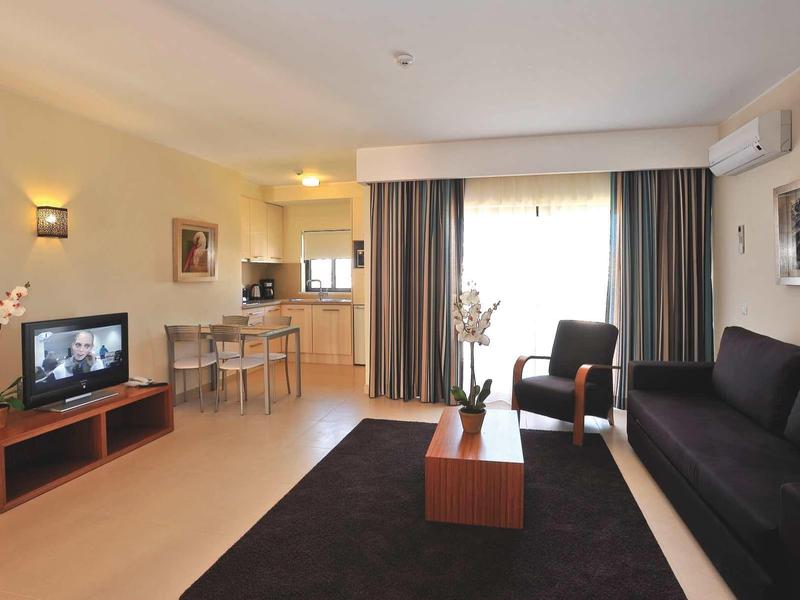 Moderner, heller Wohnraum mit schwarzem Sofa, Sessel, Fernseher, Teppich und Essbereich im Hintergrund.