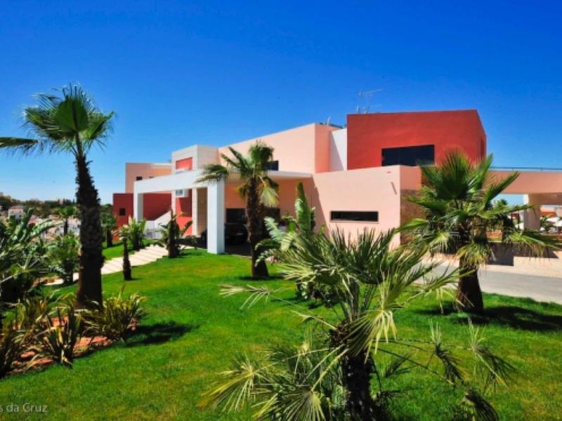 Modernes Haus mit roter und rosa Fassade, umgeben von grünen Palmen und Rasen unter blauem Himmel.