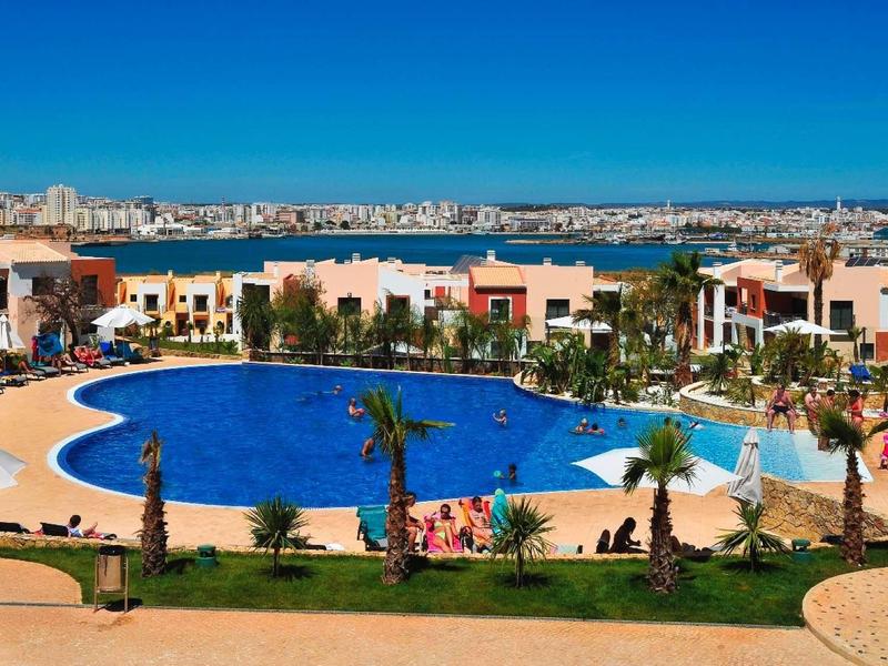 Großer Hotelpool mit Palmen, Liegen und bunten Gebäuden am Meer unter klarem Himmel.