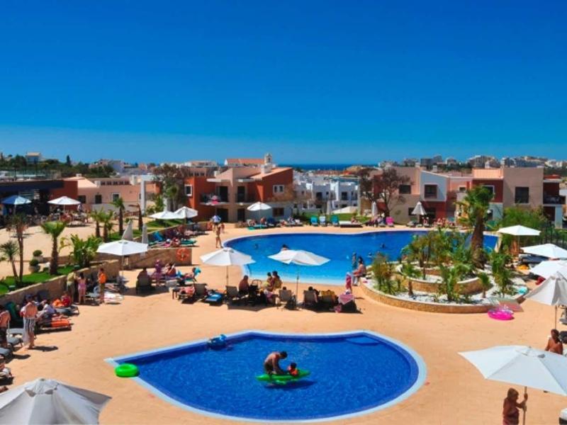 Große Hotelanlage mit mehreren Pools, Liegen, Sonnenschirmen und mediterranen Gebäuden bei klarem Himmel.