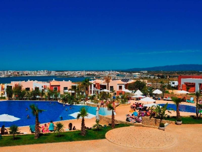 Großer Hotelpool mit Sonnenschirmen, Palmen und roten Gebäuden unter klarem blauem Himmel.
