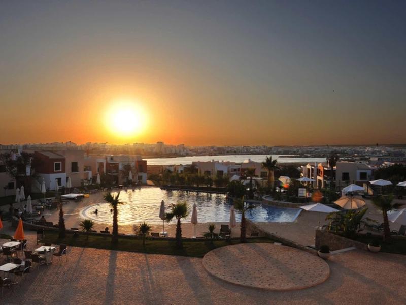Sonnenuntergang über einem Hotel mit Pool, Liegestühlen und Blick aufs Meer im Hintergrund.