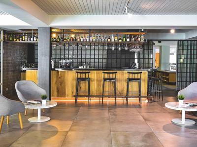 Moderner Barbereich mit Holztheke, schwarzen Hockern, grauen Sesseln und weißen Beistelltischen.