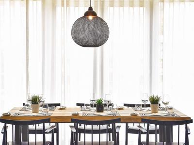 Esstisch mit schwarzen Stühlen, hellem Geschirr, Pflanzen und großer runder Hängeleuchte vor weißen Vorhängen.