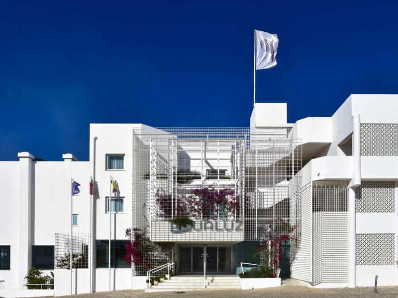 Modernes weißes Gebäude mit klaren Linien, Balkonbrüstungen und einer Flagge im blauen Himmel.