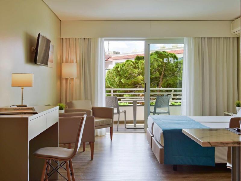 Helles Wohnzimmer mit Sessel, Tisch, Lampe und offenem Balkon mit Blick auf Bäume und Himmel.