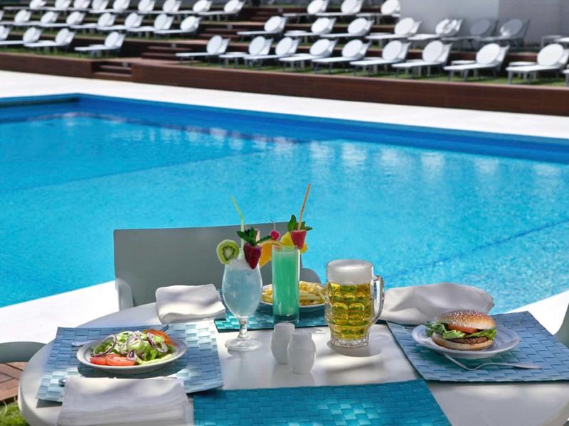 Ein gedeckter Tisch mit Getränken und Essen am Rand eines blauen Pools, im Hintergrund Sonnenliegen.