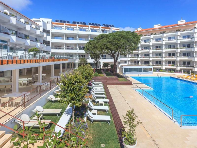 Moderner Hotelkomplex mit großem Pool, Liegestühlen, grüner Hecke und klarem blauem Himmel.