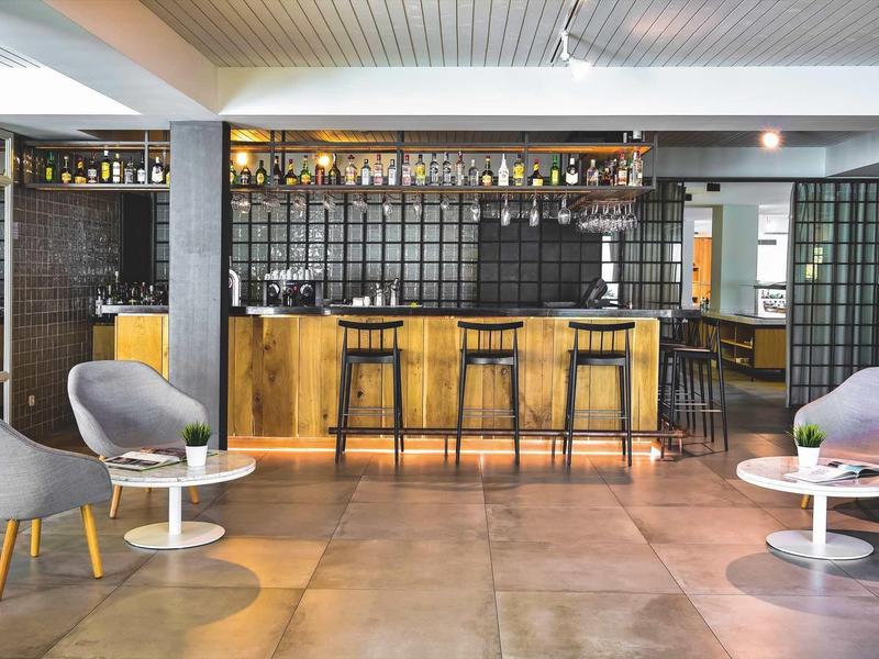 Moderner Barbereich mit Holztheke, schwarzen Hockern, grauen Sesseln und weißen Beistelltischen.