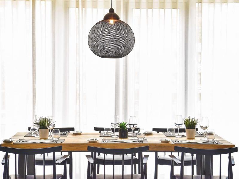 Esstisch mit schwarzen Stühlen, hellem Geschirr, Pflanzen und großer runder Hängeleuchte vor weißen Vorhängen.