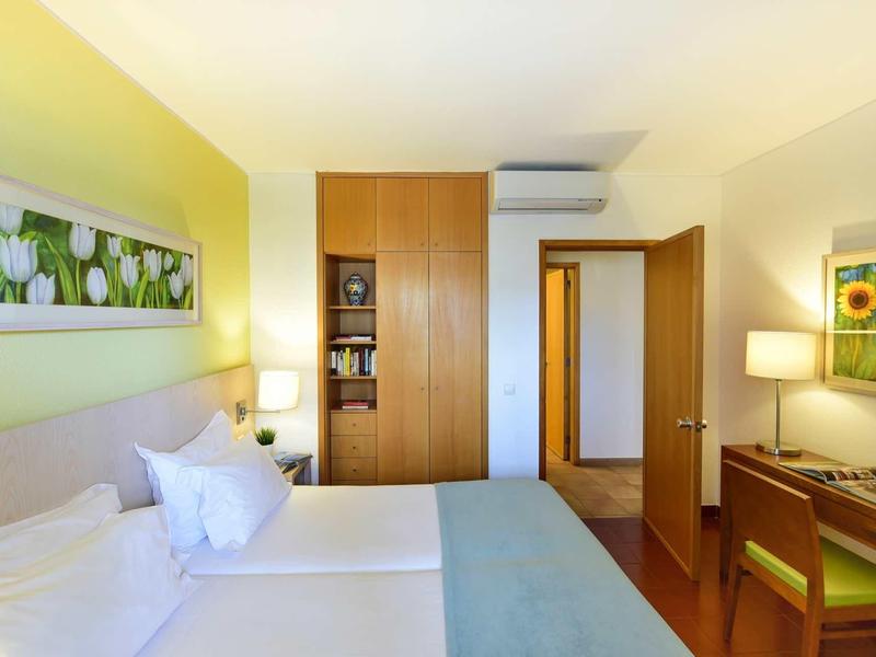 Helles Schlafzimmer mit Doppelbett, Holzschrank, Schreibtisch, Stuhl und grünem Blumenbild an der Wand.