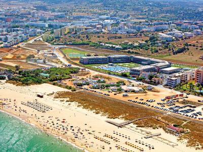 Luftaufnahme eines Küstenorts mit Sandstrand, Sonnenschirmen, Gebäuden und weitläufiger Landschaft.