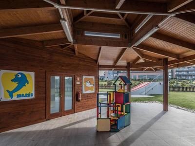 Überdachter Spielbereich mit bunter Spielstruktur und Werbeplakat an Holzwand, dahinter Grünfläche.