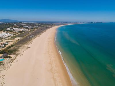 Langer, breiter Sandstrand mit klarem blau-grünem Meer und bewölktem Himmel im Hintergrund.