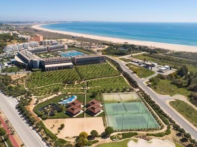 Küstenresort mit großem Schwimmbecken, grünem Tennisplatz und Sandstrand am blauen Meer.