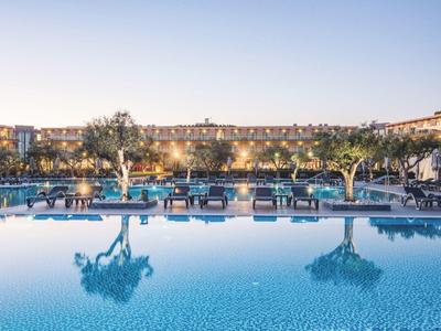 Großer, klarer Pool mit Liegestühlen und Bäumen, eingefasst von Gebäuden bei Abendlicht.