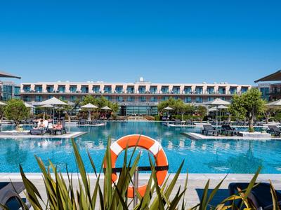 Moderner Hotelpool mit klarem Wasser, umgeben von Sonnenliegen und Gebäuden unter blauem Himmel.