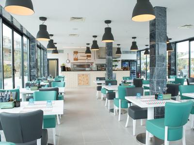 Moderner, heller Restaurantraum mit großen Fenstern, weißen Tischen und türkisfarbenen Stühlen.