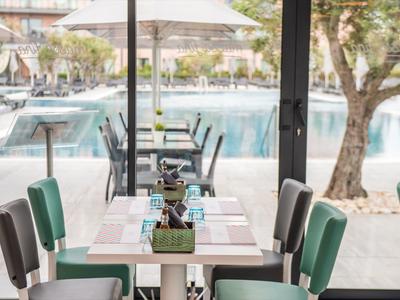 Moderner Essbereich mit weißen Tischen und grünen Stühlen, großer Glaswand und Blick auf Pool und Garten.