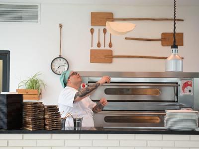 Pizzabäcker mit Bart wirft Pizzateig in einer modernen Küche über Lagertisch.