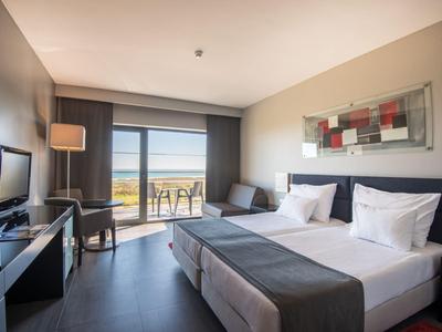 Modernes Hotelzimmer mit Doppelbett, Schreibtisch, TV, Balkon und Meerblick bei Tageslicht.