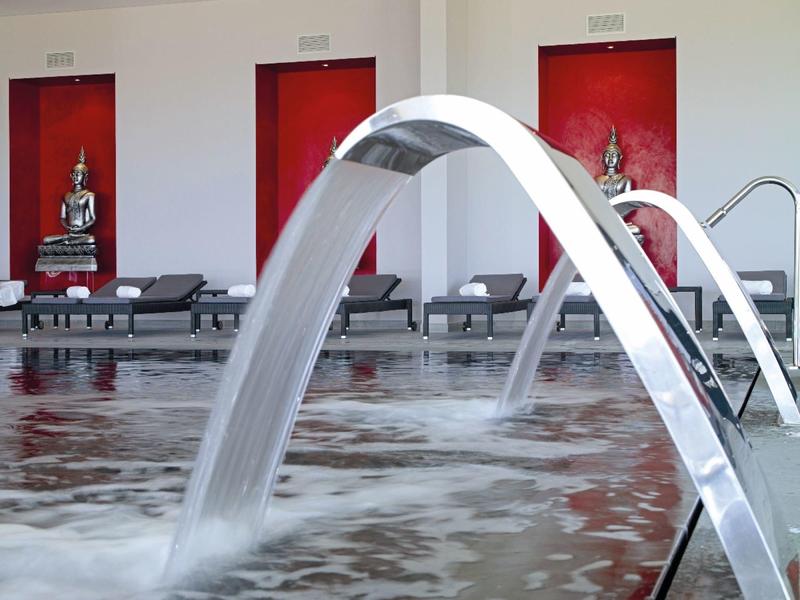 Moderne Schwimmbad-Spritzbögen aus Metall, die Wasser in ein Becken mit Sitzliegen im Hintergrund gießen.