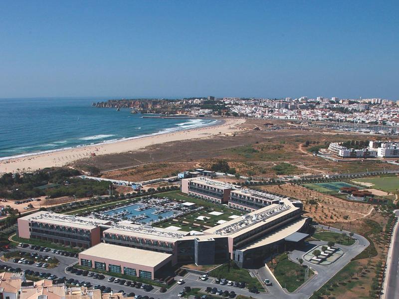 Große Hotelanlage mit Pool nahe sandigem Strand und städtischer Landschaft im Hintergrund.