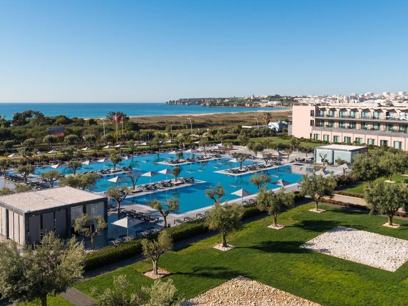 Große Hotelanlage mit Pool, Liegen, Bäumen und Blick auf Meer und Küste bei klarem Himmel.