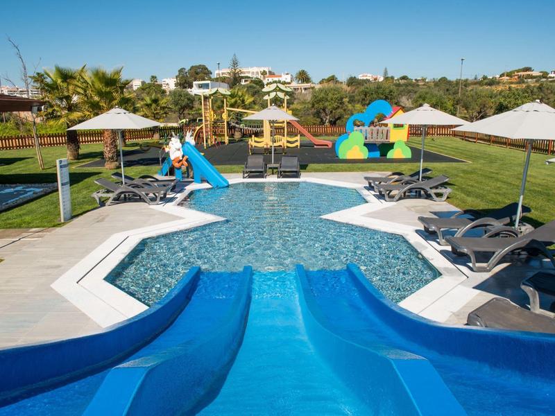 Kinderplanschbecken mit Rutsche, Sonnenschirmen und Liegestühlen, umgeben von grüner Wiese.