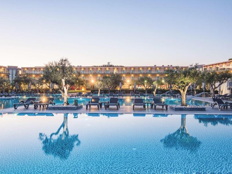 Großer, klarer Pool mit Liegestühlen und Bäumen, eingefasst von Gebäuden bei Abendlicht.