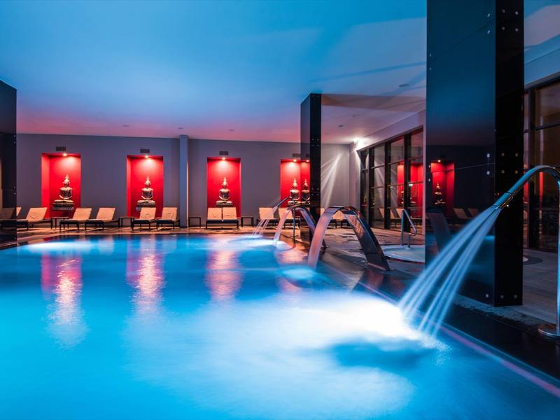 Innenpool mit blauer Beleuchtung, Wasserfall, warmem Licht und Liegestühlen an roten Wänden.