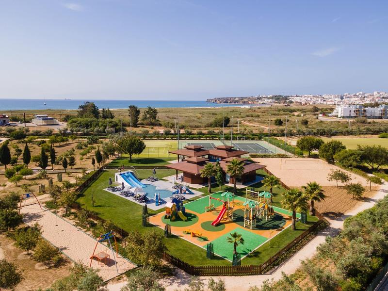 Grüner Park mit Spielplatz, Pool, Wegen und weiter Aussicht auf Felder und Häuser am Horizont.
