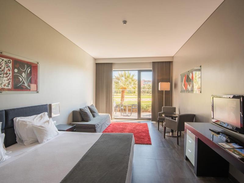 Modern eingerichtetes Hotelzimmer mit Doppelbett, Sofa, TV, Teppich und Blick auf Terrasse und Garten.