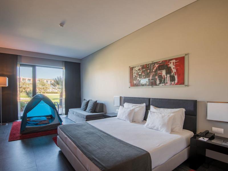 Modernes Schlafzimmer mit großem Bett, Sitzcouch, Zelt, Lampen und Balkontür mit Blick ins Grüne.